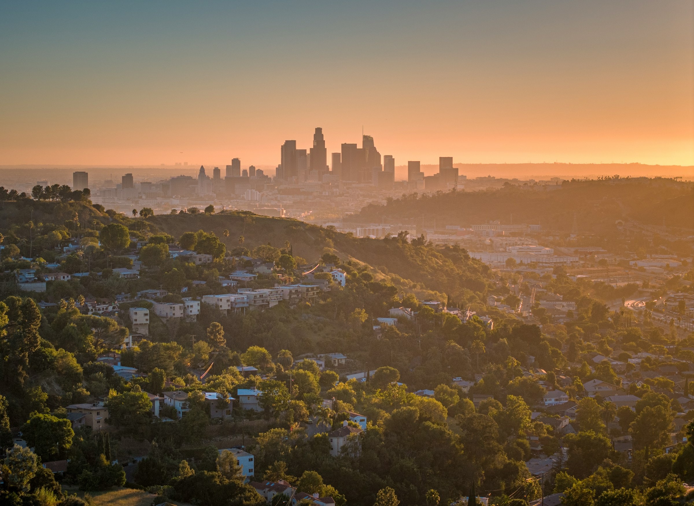 The Los Angeles skyline at sunset, viewed across hillside residential neighborhoods.
